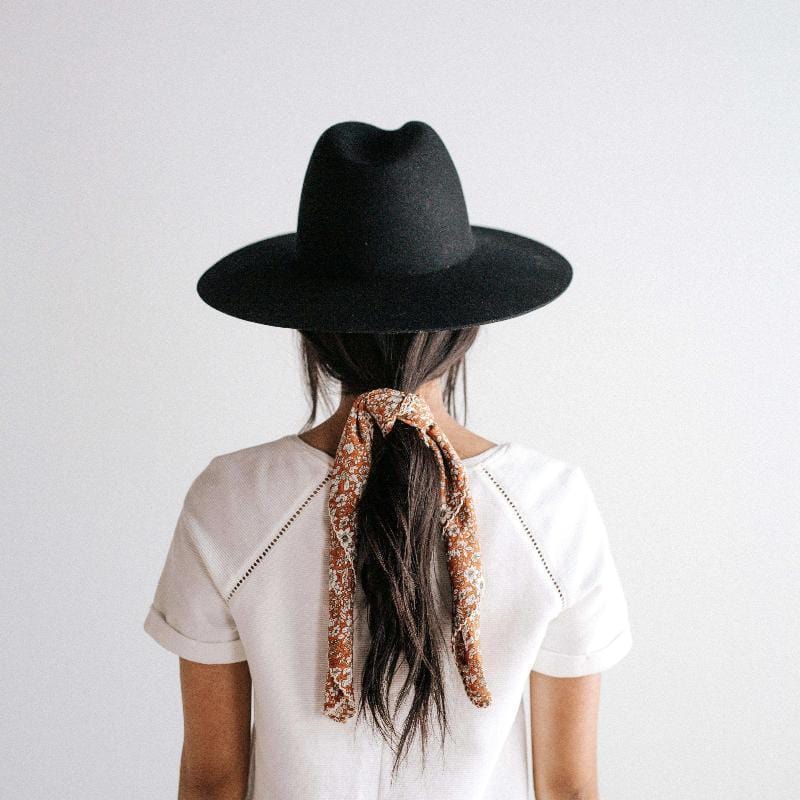 Woman wearing a black wide brim fedora hat and a white top, facing behind against a plain background #color_black