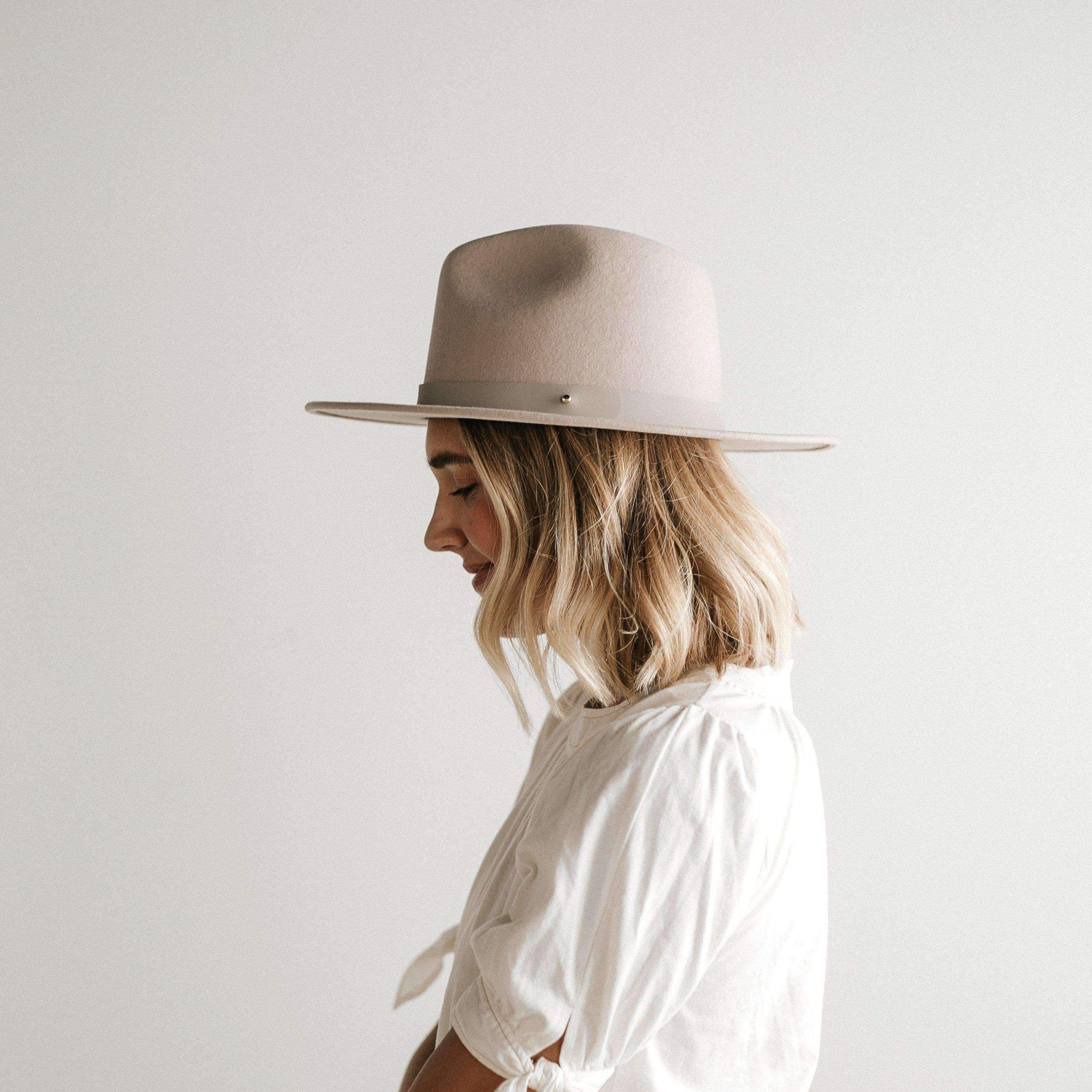 Woman wearing an ivory colored fedora hat with a hat band and a white top, facing sideways against a plain background #color_ivory