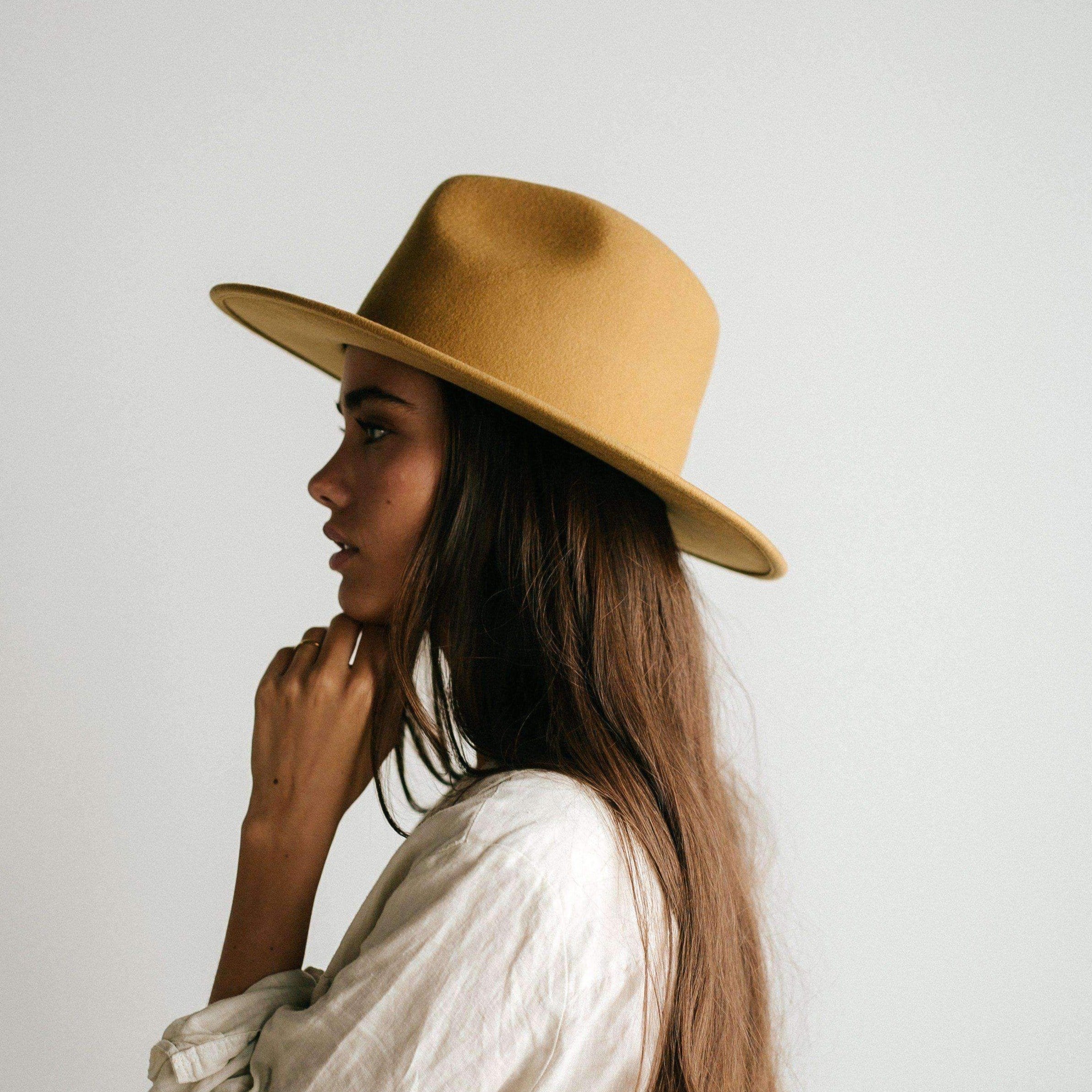 Woman wearing a mustard colored fedora hat and a white shirt, facing sideways against a plain background #color_mustard