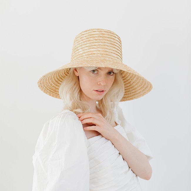 Woman wearing a natural colored boater-style straw hat and a white top against a plain background #color_natural