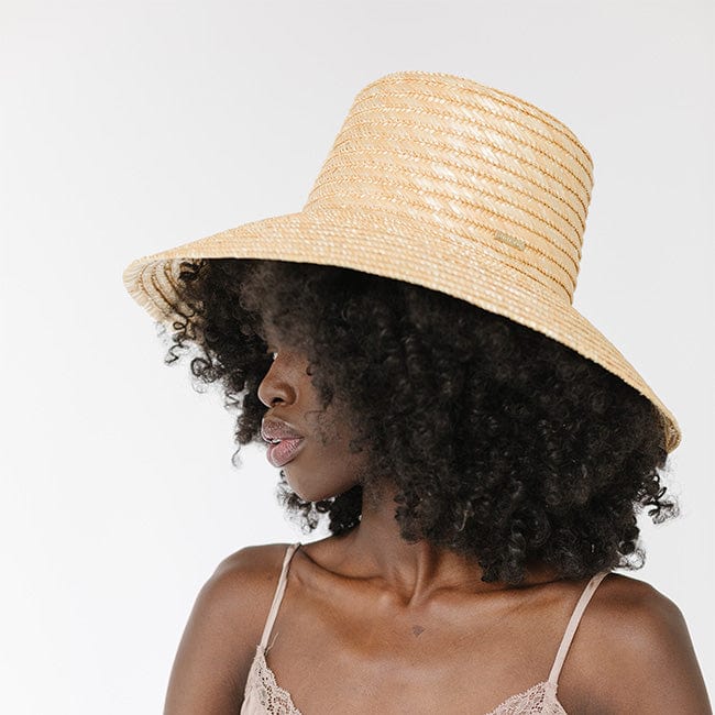 Woman with curly hair wearing a natural colored boater-style straw hat looking sideways against a plain background #color_natural