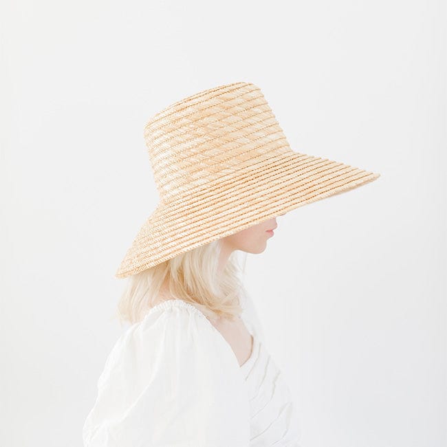 Woman wearing a natural colored boater-style straw hat and a white top, facing sideways against a plain background #color_natural